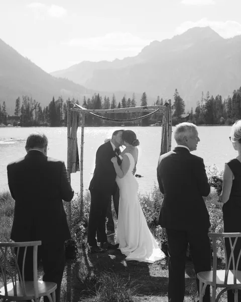 Wedding kiss as bride and groom embrace under a fabric-draped arch, bouquet in hand, with lakeside mountains and pines behind ceremony chairs