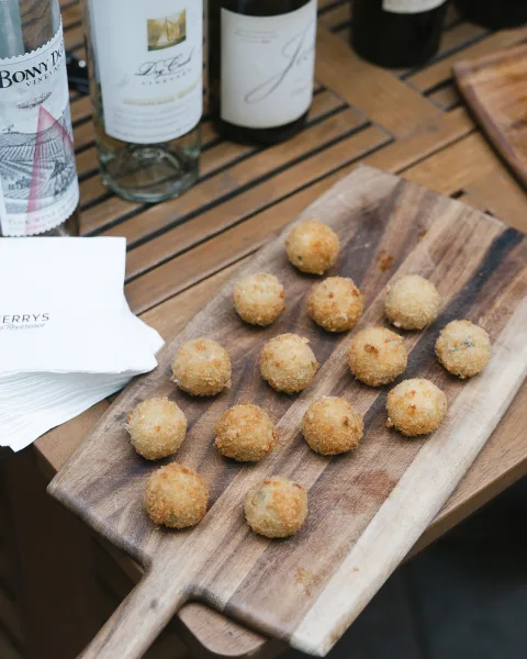 Wedding appetizers on a wooden serving board with fried bite-size croquettes and cocktail napkins beside wine bottles on a patio table