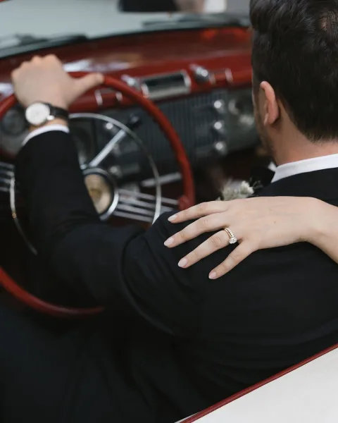 Wedding getaway car scene with groom driving in tuxedo as bride’s hand with engagement ring rests on his shoulder inside vintage interior