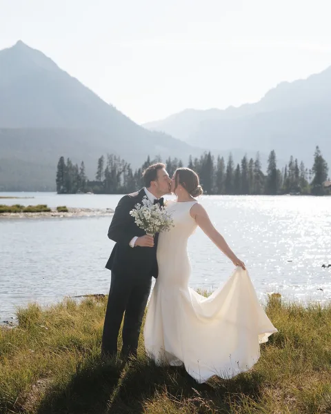 wedding kiss portrait of bride and groom kissing by a mountain lake, bride holding a white bouquet and dress train, groom in black tuxedo