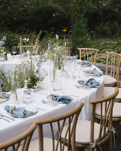 Reception tablescape with white tablecloth, blue napkin place setting, bud vases, greenery, taper candles, and wooden chairs by garden stone ledge