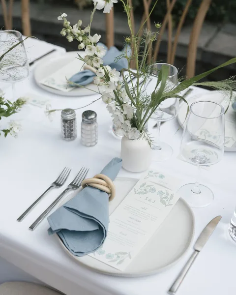 Reception tablescape with wedding place setting, blue napkin and wooden ring, printed menu, bud vase of white flowers on outdoor patio