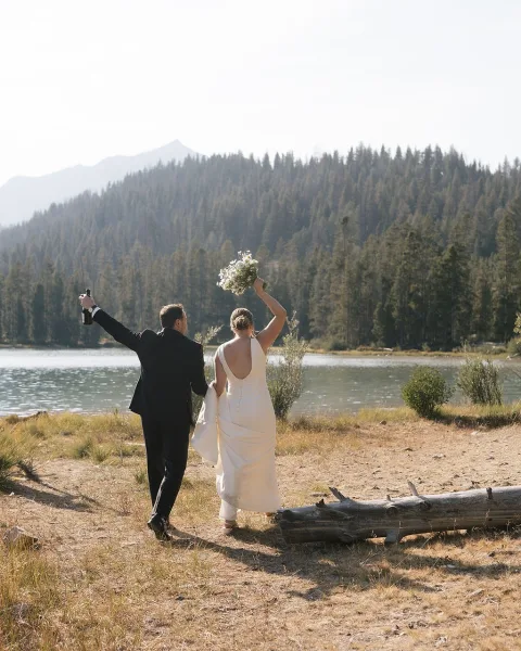 Newlywed portrait of bride and groom from behind, bride lifts bouquet as groom raises champagne bottle by a mountain lake shoreline