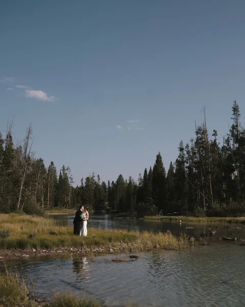 Wedding kiss portrait of bride and groom kissing, bride holding bouquet in strapless dress beside a river with pine forest and blue sky