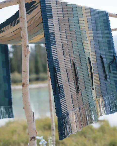 Ceremony backdrop decor with a woven wedding backdrop, fringed blue-and-white textile on birch branches by a lakeside lawn