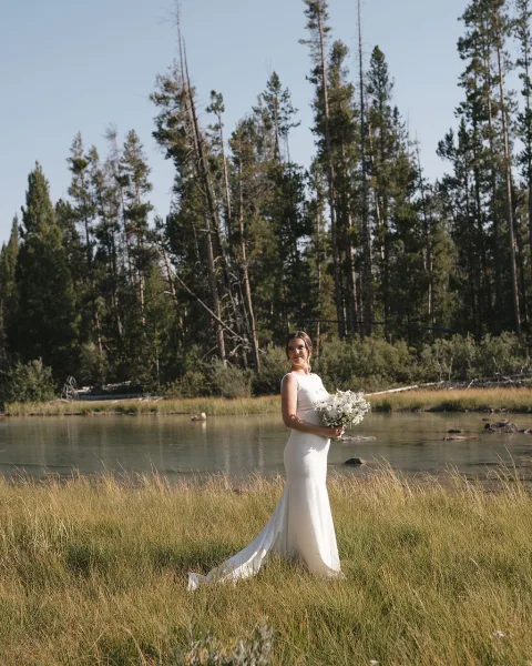 Bridal portrait of a bride holding bouquet, looking over her shoulder in a sleeveless gown by a lake meadow with pine trees and sky