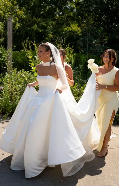 Bridal portrait of a bride in a ball gown wedding dress with cathedral veil, holding a white rose bouquet on a sunlit garden walkway