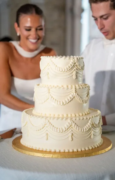 Wedding cake with white buttercream swags on a gold board, bride and groom cutting it with a knife at an indoor reception table