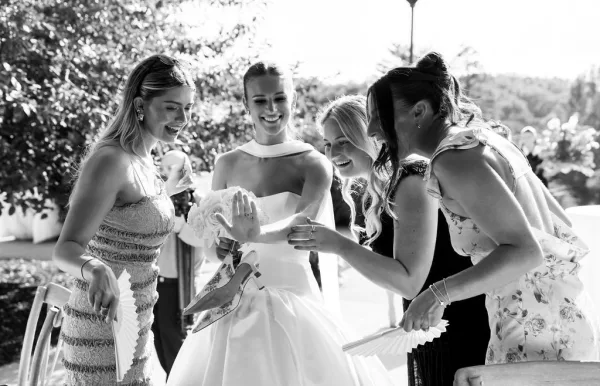 Bridal party moment as bride shows her wedding ring while bridesmaids laugh, holding bouquets on a sunny patio with trees behind