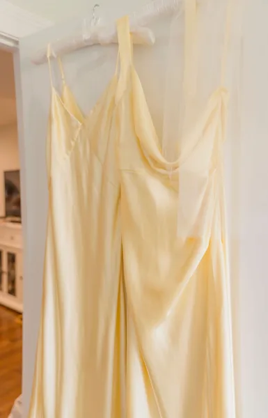Wedding dress in satin wedding dress style with spaghetti straps and cowl neckline, hanging with bridal veil in a bright room doorway
