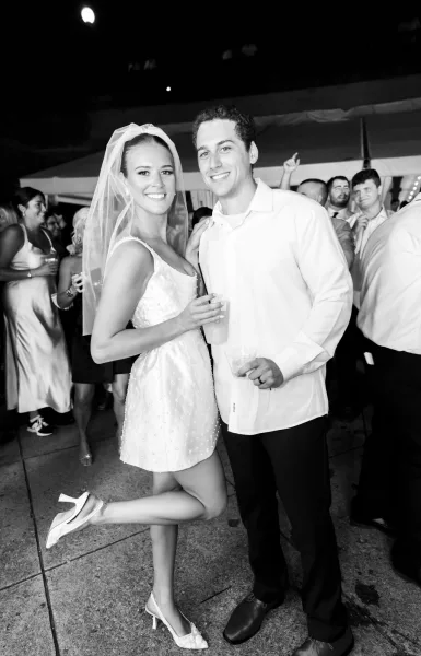 Wedding reception portrait of bride and groom smiling, holding drink cups on a tent dance floor under lights, bride in veil and heels