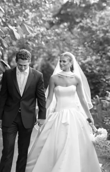Couple portrait of bride and groom walking hand in hand, her veil and rose bouquet flowing beside his tuxedo on a garden path