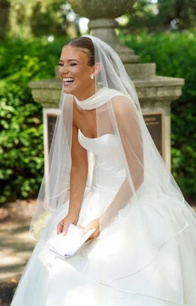 Bridal portrait of a laughing bride in a veil holding paper notes, wearing a strapless wedding dress by garden greenery and a stone fountain