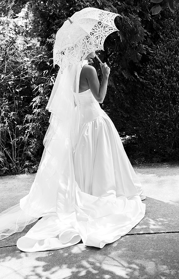 Bridal portrait of a bride holding parasol in a strapless gown with long veil and lace parasol, standing by garden trees and sidewalk