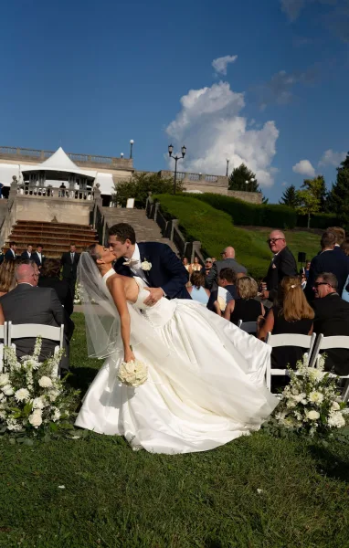 Wedding kiss as groom dips bride in strapless gown holding a white rose bouquet, guests lining a lawn aisle under blue sky