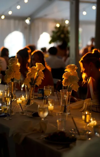 Reception tablescape with wedding table centerpieces of white roses in clear vases, votive candles and glassware under a string-lit tent with guests behind