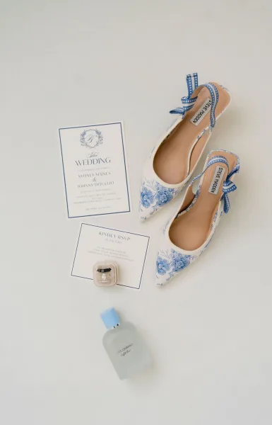 Wedding invitation suite with blue and white wedding invitations, RSVP card, rings in box, perfume, and blue floral heels on white surface
