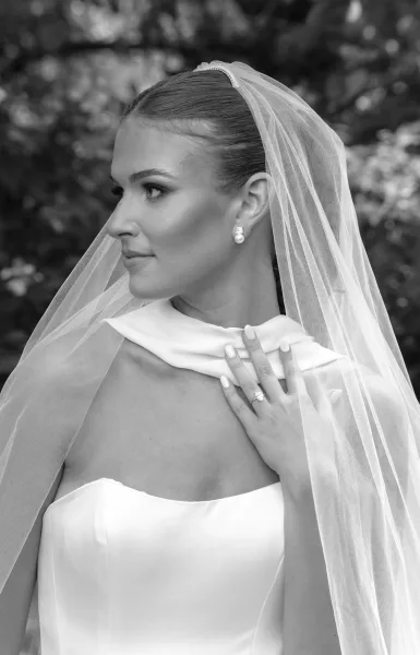 Bridal portrait in black and white of a bride in side profile wearing a strapless dress and veil, with pearl earrings and garden foliage behind
