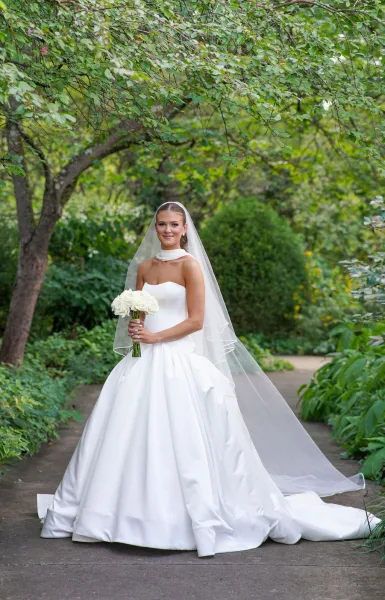 Bridal portrait of a bride in a strapless ball gown holding a white rose bouquet with a long veil trailing along a garden path