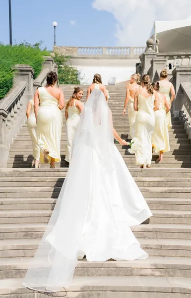 Bridal party with bride and bridesmaids in pale yellow walking up a stone staircase, bride’s cathedral veil and white bouquet trailing behind