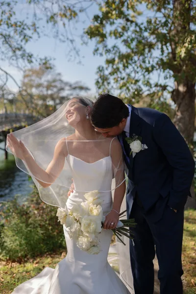 Couple portrait of bride and groom kissing as her veil blows in the wind, bride holding a white rose bouquet by a lakeside walkway.