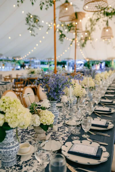 Reception tablescape with blue and white wedding table decor, chinoiserie vases, hydrangeas, taper candles, and string lights under a white tent canopy