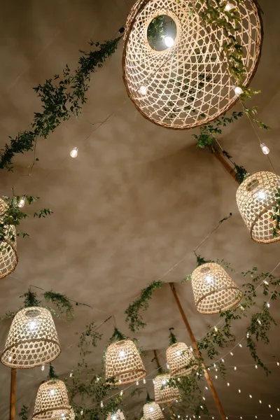 Wedding ceiling decor with rattan pendant lights and string lights, greenery garlands draped across a tent ceiling for warm ambience
