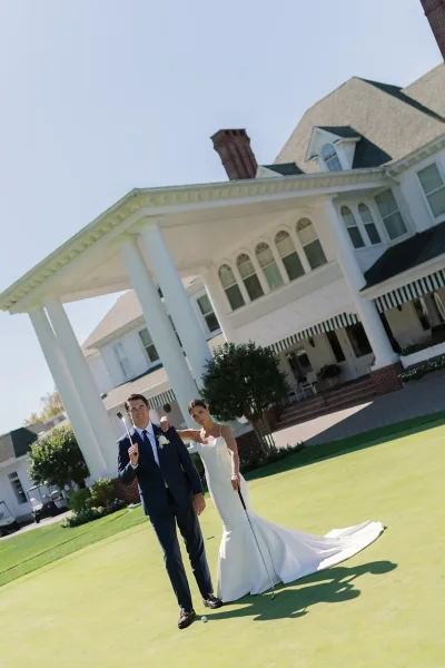 Couple portrait of bride and groom on golf course, posing with golf clubs on the green near a white-columned clubhouse under blue sky