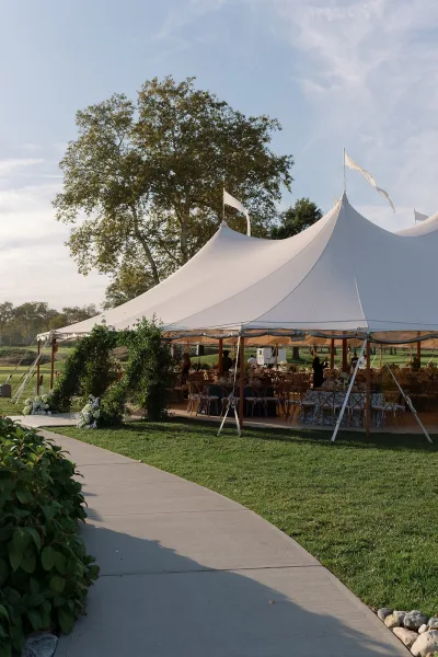 Reception tent setup beneath a white sailcloth wedding tent with flags, wooden chairs and banquet tables, framed by trees on a lawn