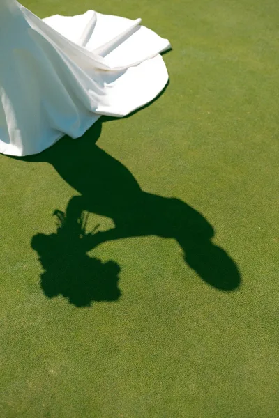Wedding dress detail with bridal dress train spread on a sunlit green lawn, bouquet in hand casting a bride shadow beside the hem