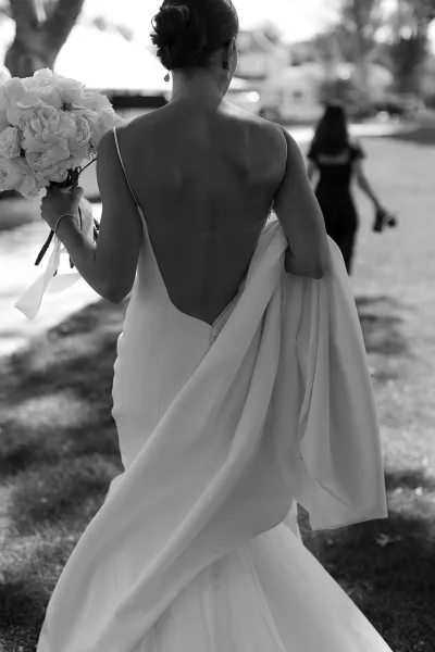 Bride portrait in a black and white wedding portrait, looking over her shoulder in a backless gown holding a ribbon-tied bouquet on a tree-lined lawn walkway