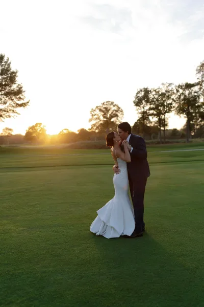 Wedding kiss portrait of bride and groom kissing on a golf course lawn at sunset, groom in navy suit and bride in strapless mermaid gown