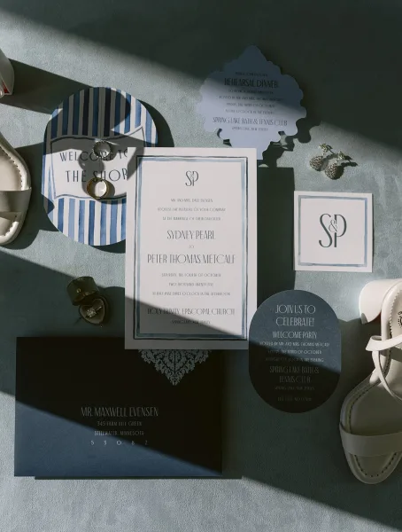 Wedding invitation suite with blue wedding invitations, RSVP and event cards, rings, earrings and white strappy heels on textured fabric in sunlight shadows