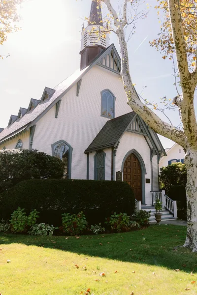 Church wedding venue with a white steeple, arched windows, and wooden doors, framed by shrubs and a sunlit lawn under blue sky
