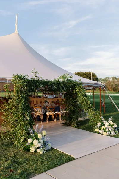 Tent wedding reception under a white sailcloth tent with a greenery arch entrance, wooden chairs, set tables, and bar on a sunny lawn