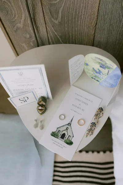 Invitation suite flat lay with ceremony program, map envelope liner, monogram card, and rings on a round side table by a wood wall