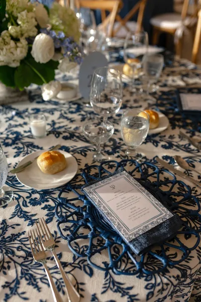 Reception tablescape with blue and white wedding table toile cloth, white and blue floral centerpiece, glass goblets, candles, and bread rolls indoors