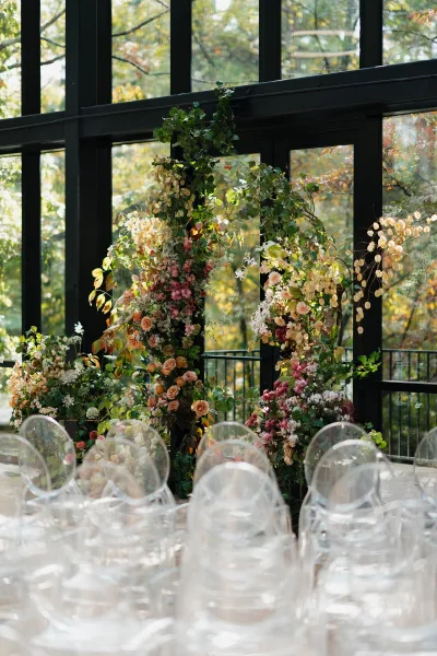 Wedding ceremony arch with an asymmetrical floral wedding arch of roses and wildflowers, hanging greenery, and clear acrylic chairs by glass windows