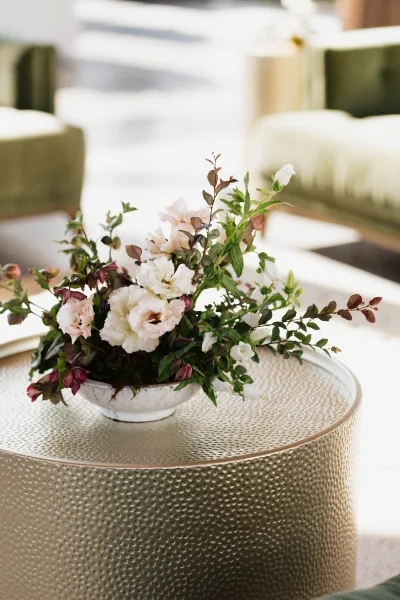 Wedding floral centerpiece in a ceramic bowl with white blooms, greenery, and burgundy foliage on a textured round coffee table in lounge seating area