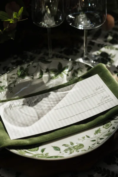 Wedding tablescape with wedding place setting featuring a printed menu card, green velvet napkin, floral plate, and wine glasses on dark linen