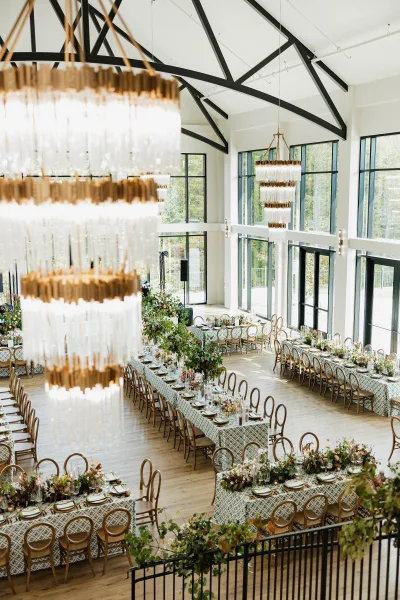 Reception tablescape with long banquet tables, taper candles and floral greenery garlands under chandeliers in a bright modern hall with tall windows