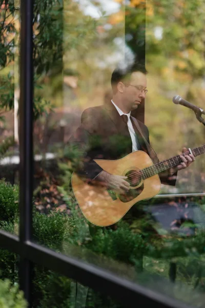 Wedding musician playing acoustic guitar at a microphone stand, wearing suit and tie with eyeglasses amid garden greenery and trees