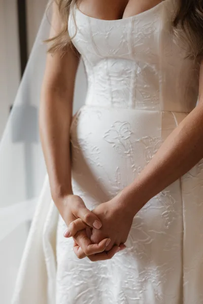 Wedding dress close-up showing a strapless wedding dress with floral applique bodice, veil drape, and bride’s nude-manicured hands clasped indoors