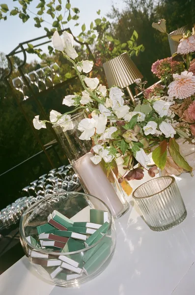Wedding welcome table decor with blush florals, tall glass candle, ribbed votive, lamp, and matchboxes on a white table in sunlight greenery