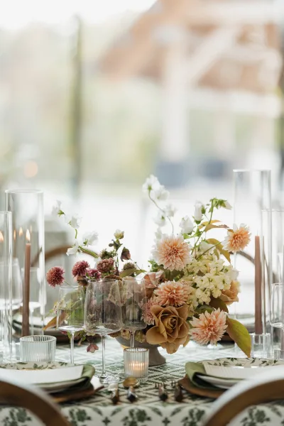 Reception tablescape with wedding floral centerpiece of blush dahlias, beige roses and greenery, taper candles in glass cylinders by bright windows