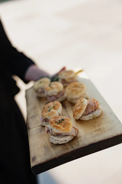Wedding appetizers on a wood serving board, biscuit slider sandwiches with sliced meat and herb garnish held by a suited server indoors