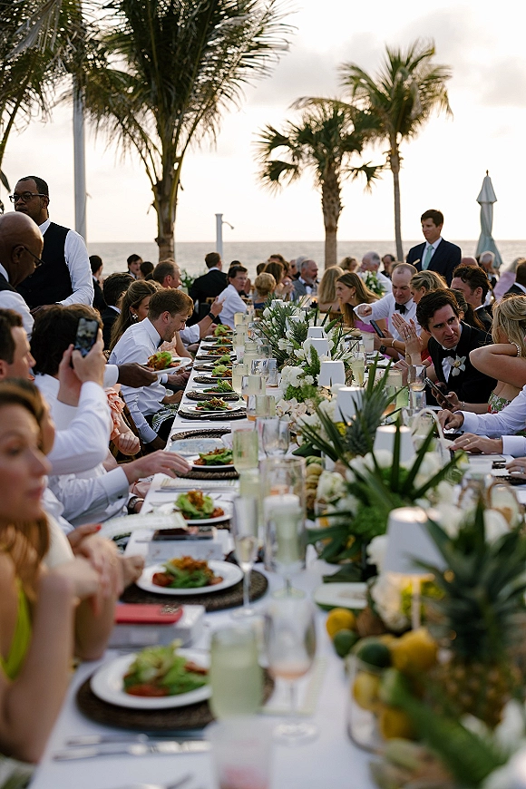 Wedding reception dinner set on a long banquet table wedding with greenery, white florals, candles, and citrus on an oceanfront patio with palms