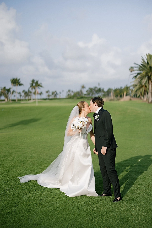 Wedding kiss portrait of bride and groom kissing, her veil and white bouquet flowing beside his black tux, on a green lawn with palm trees