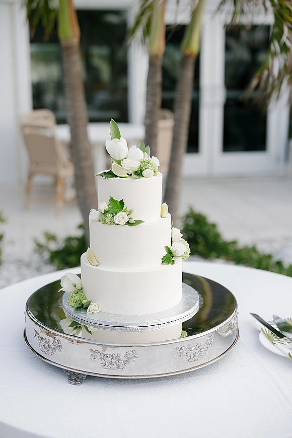 Wedding cake with three tier wedding cake design, smooth white frosting, tulips and roses, lime slices on a silver stand by palm trees