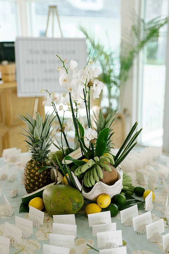 Escort card table with wedding escort cards, tropical fruit centerpiece of pineapple and citrus, and white orchids on patterned linen by large windows